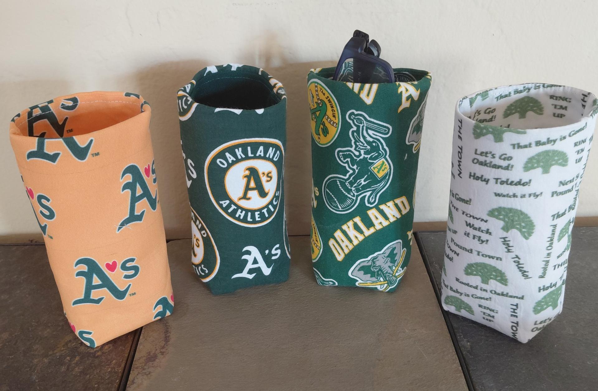 Photo of 4 different upright cylindrical upright eyeglasses holders: Gold with green As with a red heart instead of an apostrophe; green with circular A's logo; green with all over scattered A's logos, elephants and the word Oakland; and white with various A's words and sayings.
